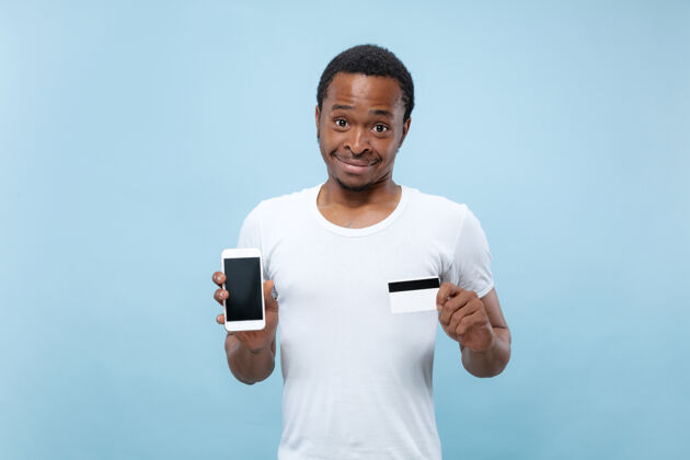 选择在蓝色空间里 一个身穿白衬衫 手持卡片和智能手机的年轻非裔美国人的半身肖像成功在线广告