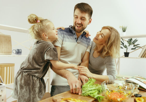 晚餐可爱的小女孩和她美丽的父母在家的厨房里切蔬菜 一边微笑着做沙拉吃爸爸健康