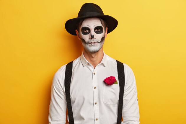 魔鬼严肃的男性僵尸的肖像戴着头盖骨面具 化着可怕的妆 庆祝墨西哥的节日 戴着黑色帽子和白色衬衫 带吊带 口袋里有红玫瑰 黄色背景下是孤立的头骨邪恶男性