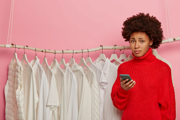 时尚不满悲伤的非洲发型女人拿着手机 站在衣柜旁边 手里拿着手机摆姿势 心烦没有时髦的服装年轻电话红色