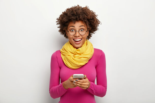 快乐热情有趣的卷发年轻女子手持现代手机 阅读短信 戴着眼镜和粉色高领毛衣 在白色背景下摆姿势科技概念年轻时尚室内