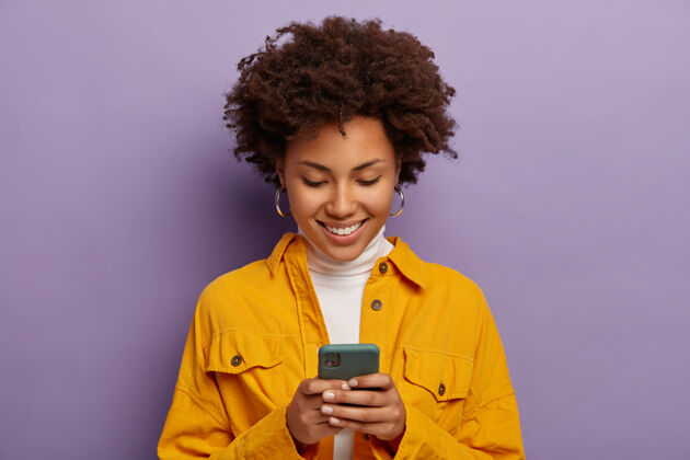 黄色快乐女性的照片 专注于智能手机 穿着黄色衬衫 微笑温柔 孤立在紫色的工作室墙上室内女人手机