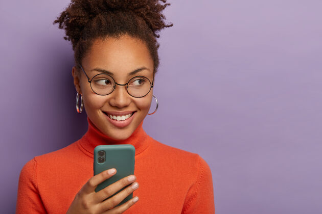 电话积极漂亮的非裔美国年轻女子专注在一旁 戴着圆眼镜和休闲套头衫 开心地笑着阅读眼镜黑色