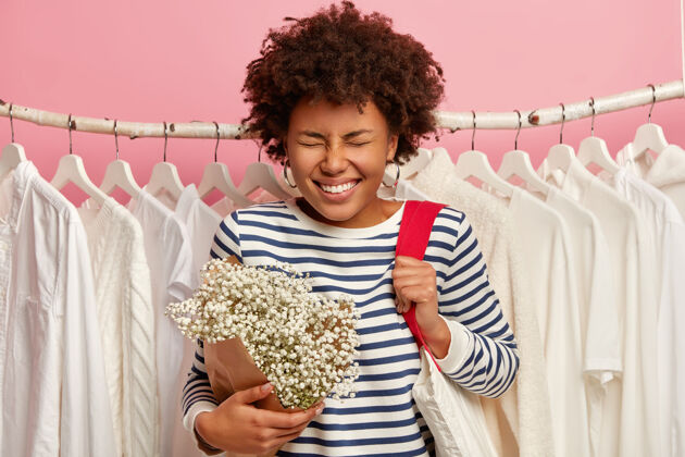 悬挂快乐的非洲发型女人 开心地笑着 拿着购物袋 漂亮的花 站在衣柜里的白色衣服的衣架上 很高兴来到时装店情感和销售理念非洲商店高兴