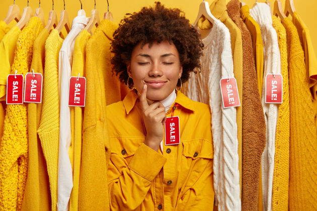 夹克一张可爱的非洲发型女人的照片 在服装店试穿新的黄色夹克 闭上眼睛 站在有红色标签的衣服中间 寻找时髦的服装Consumer室内Glad
