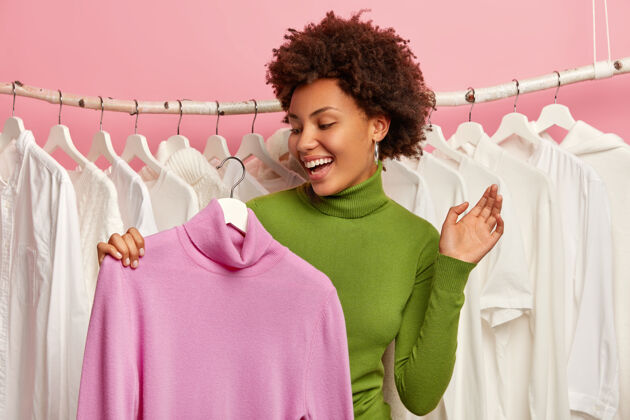 黑发年轻的非洲女人很高兴找到了一件套头衫 把紫色的高领毛衣挂在衣架上 为聚会挑选服装 在精品店购物 保持手掌抬高购买欢乐粉色