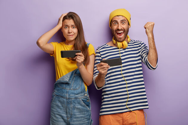 年轻年轻的女人和男人在智能手机上玩电子游戏 男人展示拳头碰撞 庆祝胜利 穿着条纹套头衫和耳机 担心的女人站在附近紫色男人站着