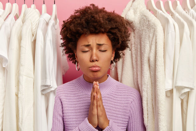 服装冷静的卷发女人双手合十 祈祷幸福 穿着针织毛衣 站在衣架上的白色衣服之间 在粉色背景上与世隔绝紫色紫色民族