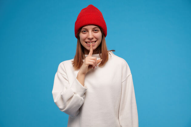 服装有吸引力的 深色头发的成年女孩的肖像穿着白色毛衣和红色帽子显示沉默的标志和微笑隔离在蓝色的墙上黑发孤独室内