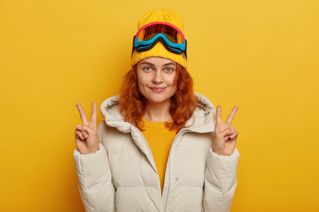 面具半身镜头的姜美女举起双臂 做出和平或胜利的手势 用滑雪镜 穿着冬天的白色外套隔离在黄色背景上红发背心冬天