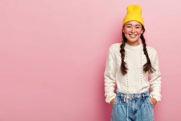 脸快乐的亚洲女人带着温柔的微笑 双手插在牛仔裤的口袋里 戴着黄色的帽子 白色的套头衫 在玫瑰色的墙壁空白处摆着两个辫子的姿势眼睛冷女人
