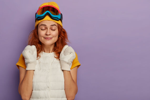 红发运动型的年轻女子喜欢滑雪场 胜利地握紧拳头 闭上眼睛 戴着保护性的滑雪板眼镜 在紫色的工作室墙上与世隔绝女士室内请
