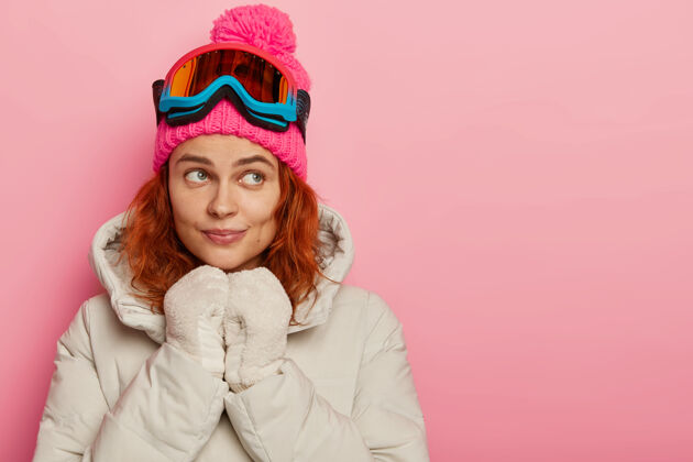 梦幻体贴的女性化女孩 姜黄色卷发 戴着暖和的帽子 外套和手套 戴着防护性的滑雪眼镜 在粉色背景下在室内摆姿势思考一边护目镜