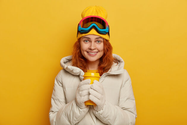 面具女滑雪者穿着温暖的冬季外套 拿着黄色的外卖杯和热茶 戴着帽子和滑雪镜 愉快地微笑 在室内做模特饮料微笑服装