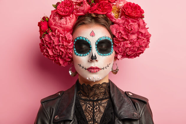 花用传统墨西哥脸谱画的美女特写肖像 闭着眼睛 戴着芳香的花朵做的花环 黑色的衣服 在粉红色的墙上摆姿势深色装饰油漆
