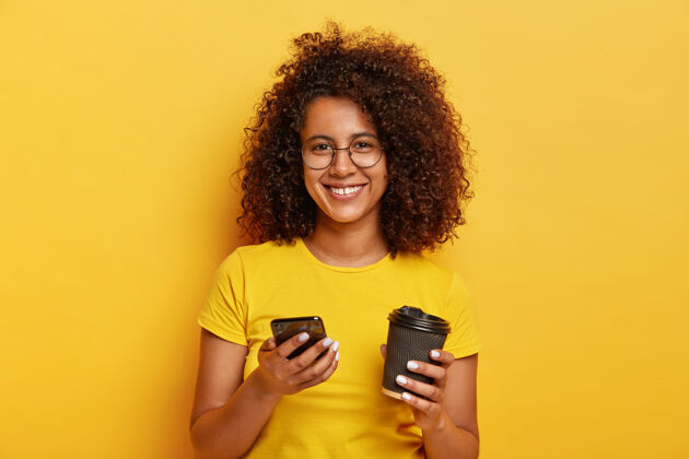 手机可爱的千禧一代女性肖像手持智能手机喝外卖咖啡 笑容可掬 身穿黄色t恤 请号码稍后见面T恤单色站立