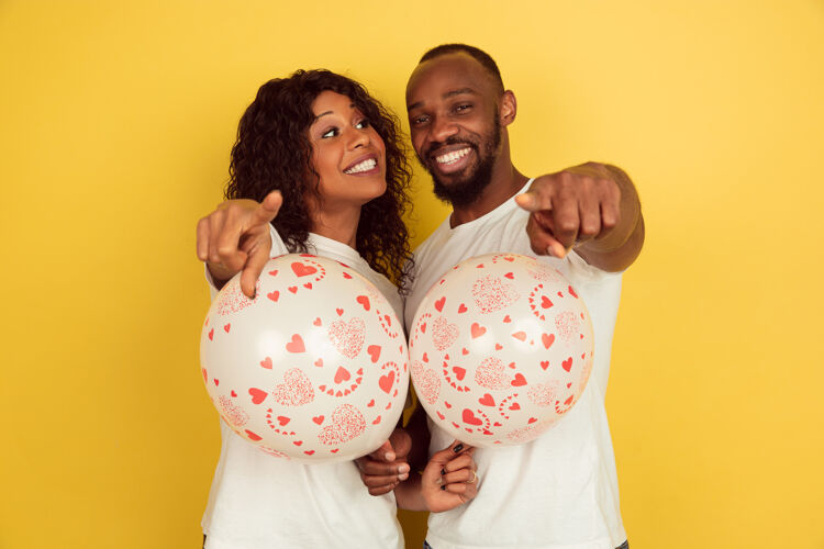 男性指着气球情人节庆典 黄色背景上的一对幸福的非洲裔美国夫妇关系微笑两个