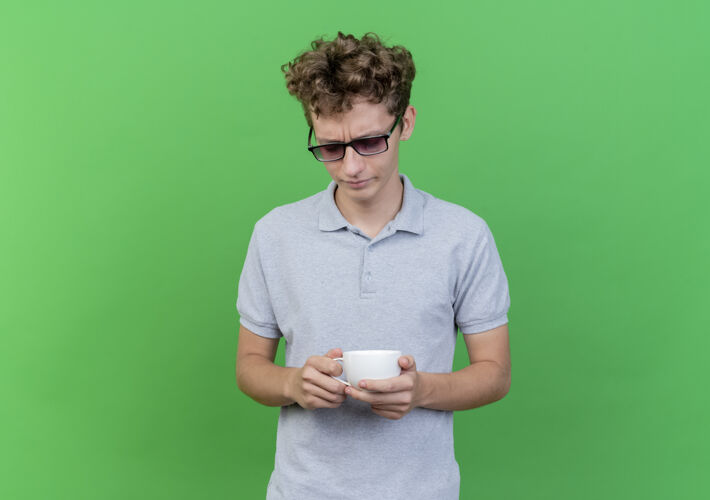 男人一个戴着黑眼镜 穿着灰色马球衫 拿着咖啡杯的年轻人站在绿色的墙上 神情悲伤地低头看着戴着年轻放下