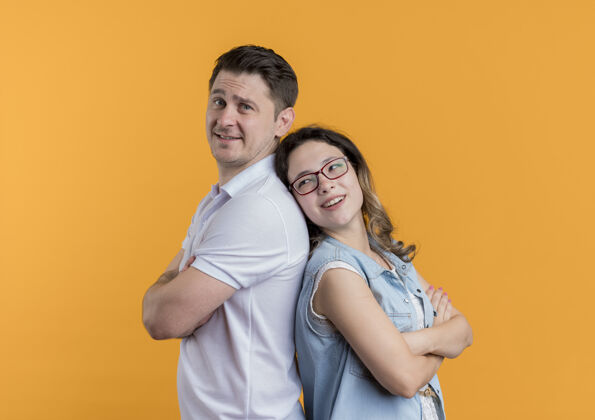 衣服一对穿着休闲服的年轻夫妇背靠背地站在一起 幸福而积极地微笑着站在橙色的墙上微笑站立男人