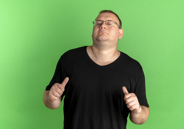 指着一个戴眼镜 身穿黑色t恤的胖男人站在绿色的墙上指着自己戴他自己超重