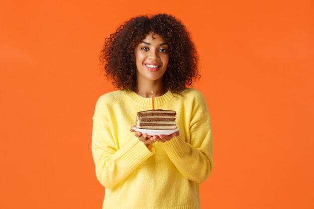 脸腰部向上的肖像画美籍非洲裔美籍b日女孩 留着非洲式发型 拿着生日蛋糕的盘子 点燃蜡烛 吹着它许愿 开心地微笑 在橙色的墙上庆祝蛋糕休闲糕点