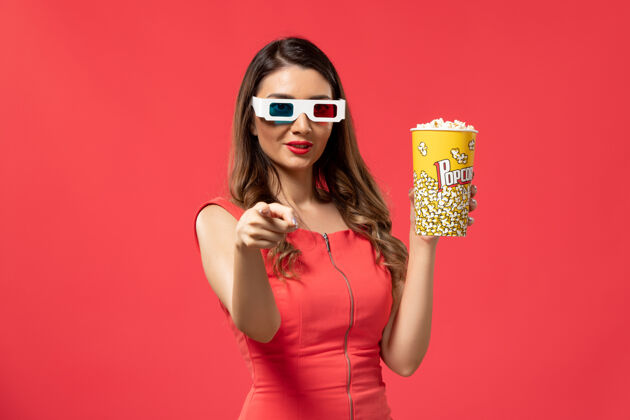 女士正面图：戴着d型太阳镜拿着爆米花的年轻女性在浅红色的表面上微笑电影太阳镜女性