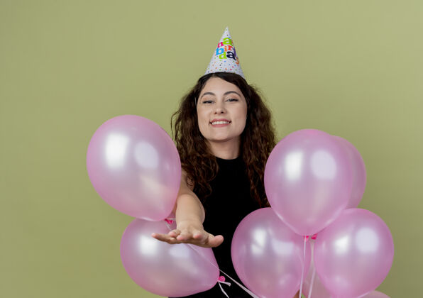 空气年轻漂亮的女人 卷发 戴着节日帽 手持气球 欢庆生日派对快乐而积极的微笑 欢快地站在明亮的墙上庆祝年轻积极