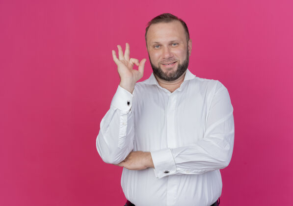 自信一个留着胡子的男人穿着白衬衫 微笑着自信地站在粉色的墙上 展示着ok标志男人胡须站