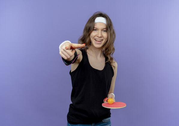 网球身穿黑色运动服的年轻健身女孩 头上戴着头带 手持球拍和乒乓球 食指微笑着站在蓝色的墙上桌子微笑头带