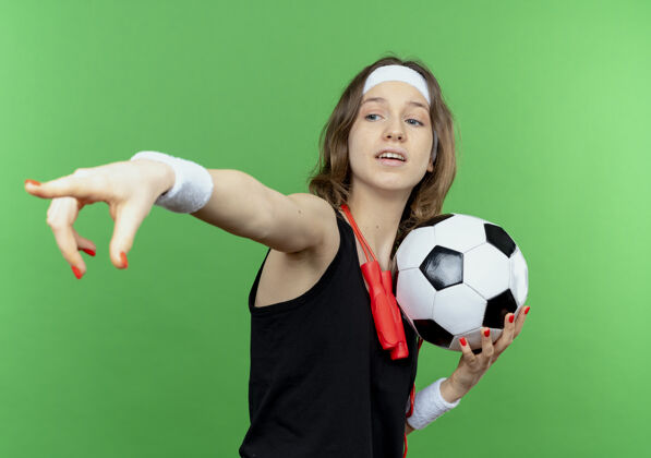 女孩穿着黑色运动服的年轻健身女孩戴着头带 脖子上系着跳绳 手里拿着足球 手指指着绿色的墙上的东西健身跳过绳子