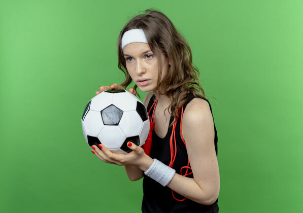 抱身穿黑色运动服 头戴头带 脖子上系着跳绳 手持足球 表情严肃的年轻健身女孩站在绿色的墙上绳子跳过女孩