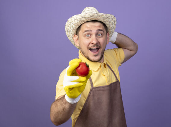 工作年轻的园丁 穿着连体衣 戴着帽子 戴着工作手套 手里拿着新鲜的西红柿 开心而积极地笑着连身衣欢呼年轻