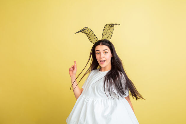 肖像指着 微笑着白人妇女作为一个复活节兔子在黄色工作室背景复活节快乐的问候情感姜黄色表情