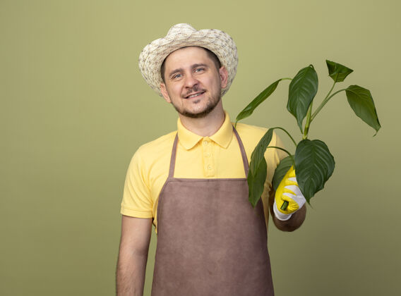 植物年轻的园丁 穿着连体衣 戴着帽子 戴着工作手套 面带微笑地拿着植物连身衣年轻人男人