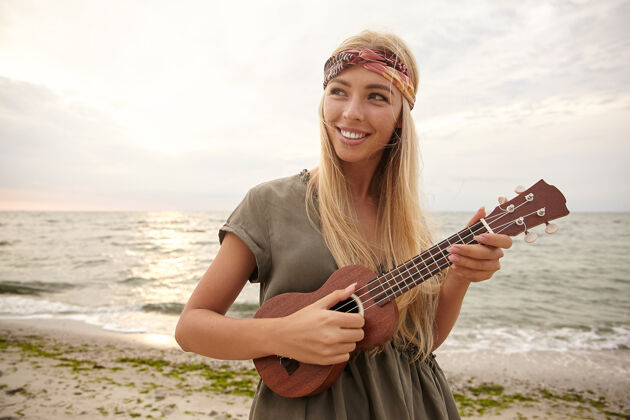 绿色年轻迷人的白头女子戴着头巾 一边快乐地笑着 一边弹着小吉他 在明媚温暖的日子里漫步在海边的户外照片海滩女性请