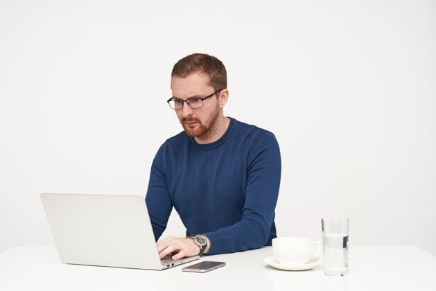 毛衣室内镜头：戴着眼镜 留着胡须的年轻人一边认真地看着笔记本电脑的屏幕 一边工作 一边把手放在键盘上 与白色背景隔离开来年轻人水姿势