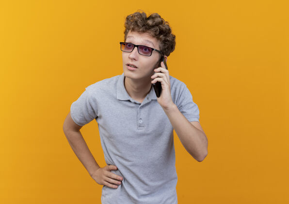 惊喜一个戴着黑眼镜 穿着灰色马球衫的年轻人站在橘色的墙上讲着手机 看上去很惊讶男人电话眼镜