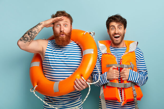 男性快乐的家伙们穿着救生衣和救生圈在海滩上摆姿势水手姿势在一起