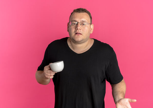 眼镜一个戴眼镜的超重男人 穿着黑色t恤 拿着咖啡杯 耸耸肩 站在粉红色的墙上 看起来很困惑杯子拿着咖啡