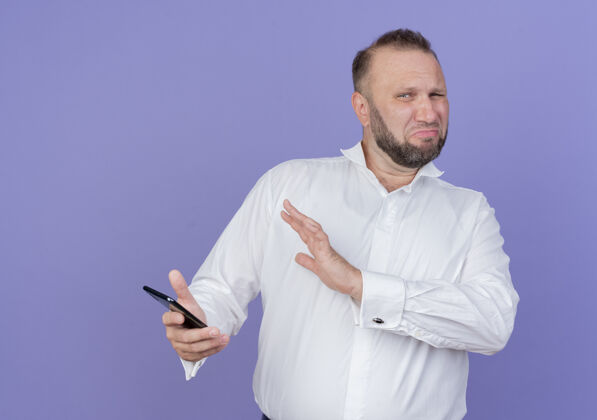 表情一个留着胡子的男人穿着白衬衫 拿着智能手机 站在蓝色的墙上 用手做着防御的手势 带着厌恶的表情男人握着防御