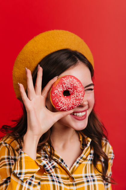 可爱积极的卷发女孩眨眼 用草莓甜甜圈遮住眼睛穿着格子衬衫 戴着黄色帽子的漂亮女人在红墙上摆姿势黑发女孩法国