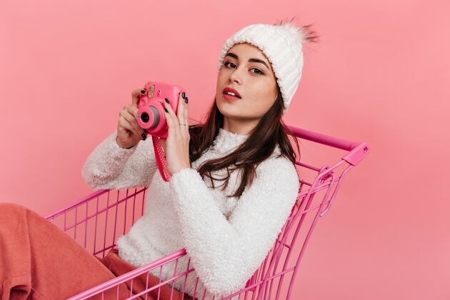 享受一个可爱的女孩 穿着白色冬衣 手里拿着粉红色的相机 坐在超市的手推车里 在孤零零的墙上摆姿势白色毛衣年轻肖像