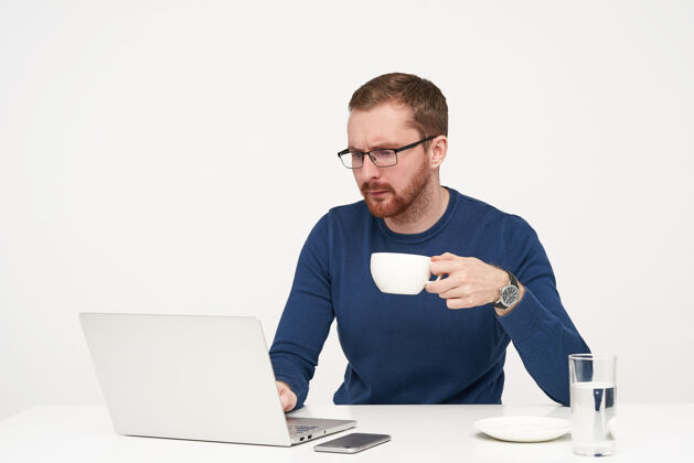 理发严肃的年轻帅哥 留着胡子 高举着一杯茶 看着笔记本电脑的屏幕 一张专注的脸 隔离在白色的背景下年轻人手表水