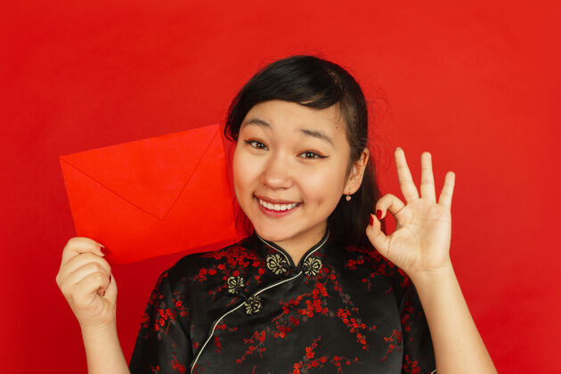 新年中国新年红色背景上孤立的亚洲少女肖像穿着传统服装的女模特特写看起来很开心 并显示红包庆祝 节日 情感亚洲年轻装饰