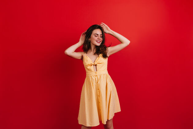 五颜六色黑发好幽默的女孩在红墙上悠闲地摆着姿势穿着鲜艳夏装的女人微笑着喜悦黄色连衣裙服装