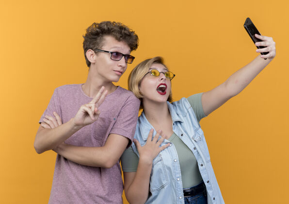 站立年轻漂亮的夫妇站在一起用手机自拍微笑着在橙色的墙上展示v形标志情侣微笑使用