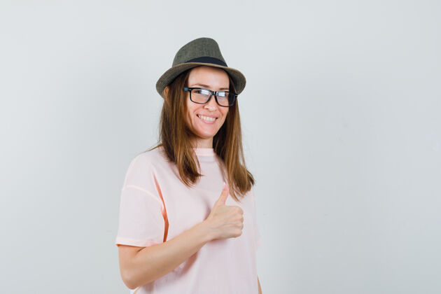 肖像年轻女孩展示拇指在粉红色的t恤衫 帽子和看起来愉快 前视图快乐帽子微笑