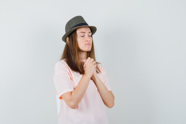 帽子年轻的女性在祈祷中双手合十 穿着粉色t恤 戴着帽子 看上去很平静前视图时尚积极漂亮