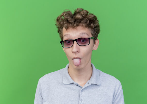 马球一个戴着黑色眼镜 穿着灰色马球衫的有趣的年轻人站在绿色的墙上做鬼脸 伸出舌头看鬼脸年轻