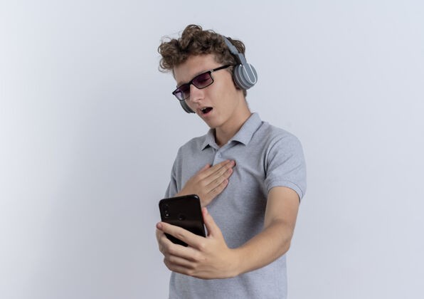 穿一个戴着黑眼镜的年轻人 穿着灰色马球衫 戴着耳机 站在白墙上看着他的智能手机屏幕 心怀感激耳机屏幕男人